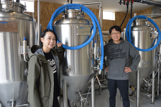ビールの醸造所の設備の傍らに立つ大輪真理子さんと夫の弘詳さん