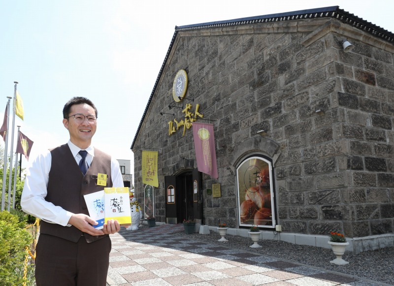 石造りの「The Sun蔵人」の店舗外観と、その前で商品を手にする支配人の西川洋平さん