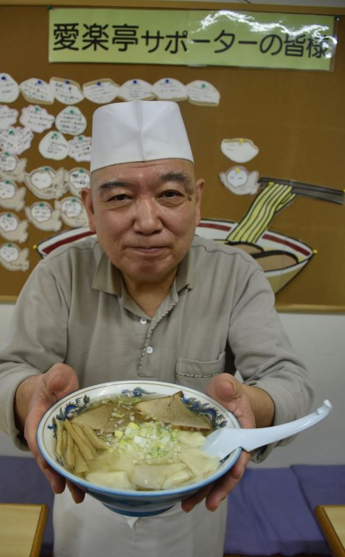 店内に掲示されている「愛楽亭応援券」の前で「たくさんの応援に感謝しかない」と話す店主の木戸日出明さん