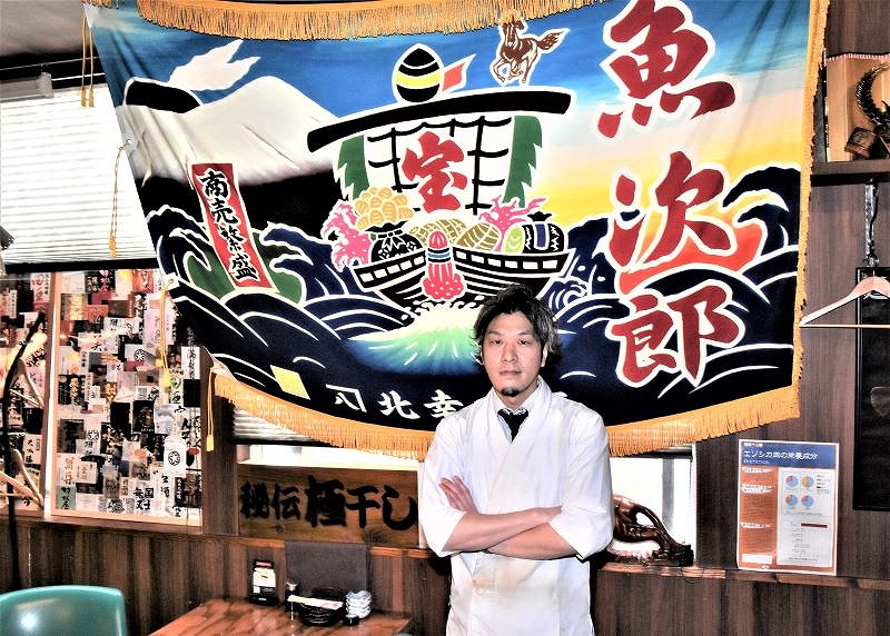 店内に飾られた常連客からの贈り物の大漁旗を前に立つ店長の袖村亮太さん