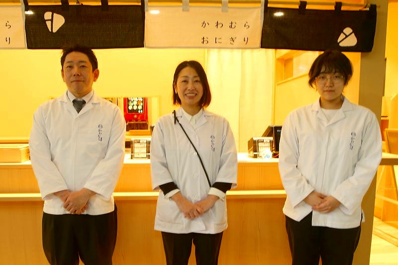 「かわむら出汁茶」のおにぎり屋さんの店舗前で笑顔を見せる３人のスタッフ