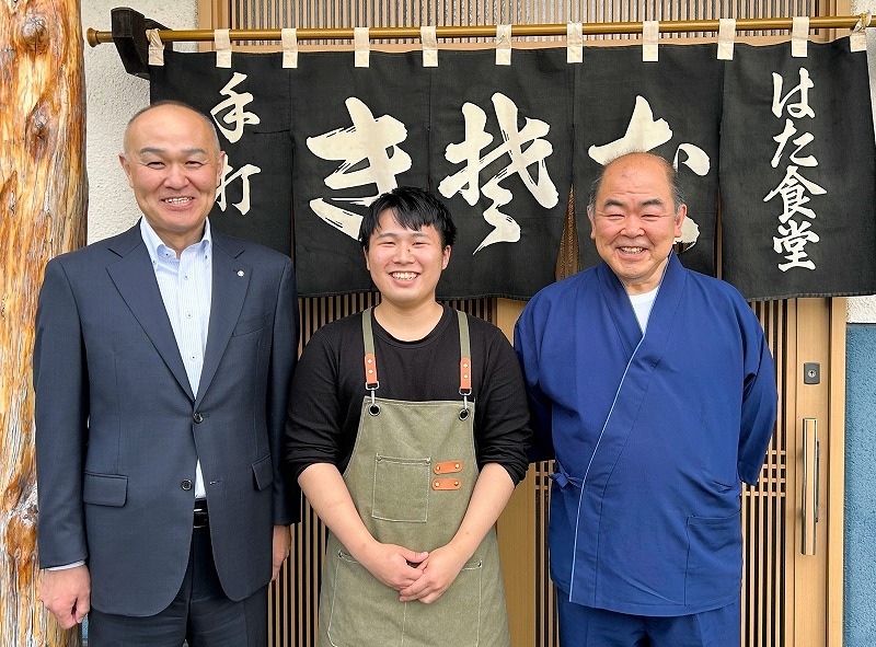 そば店「秦食堂」の秦代表（右）や本田町長（左）と店前に並び、笑顔を見せる山崎さん