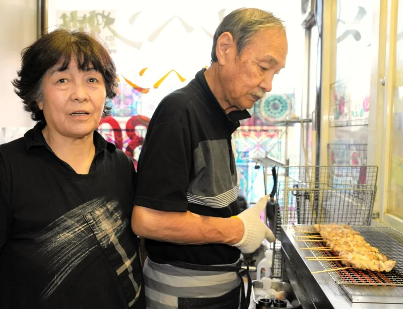 「駄菓子と一緒に親のおつかいで焼き鳥を買う子もいる」と話す店主の川原正子さん（左）と、焼き鳥を焼く夫の直幸さん