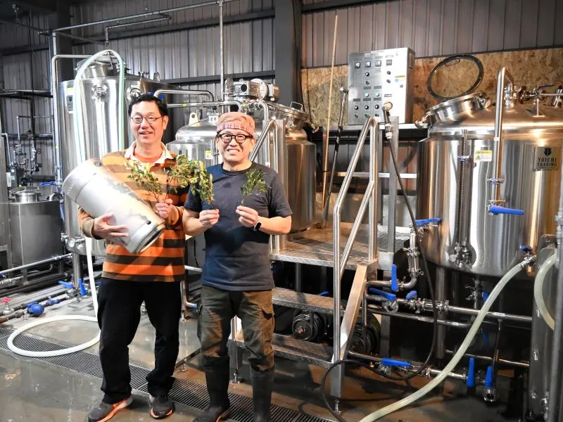 念願のクラフトビール醸造所の稼働に笑顔を見せる高橋さん（右）と北海道ブルワリー関係者。手に持つのは原料のヤチヤナギ