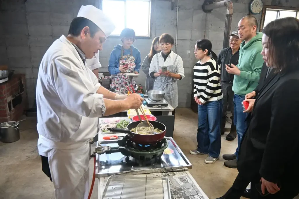 天ぷらを揚げる成田さん(左)を見つめる長谷川さん(右から5人目)やモニターツアーの参加者ら